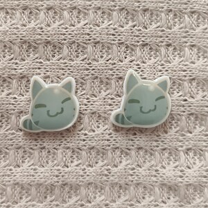 May include: Two light blue cat-shaped pins with smiling faces and striped tails. The pins are set against a textured, off-white fabric background. The cat pins are a cute accessory.