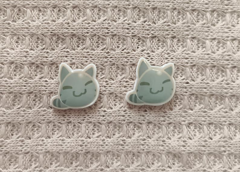 May include: Two light blue cat-shaped pins with smiling faces and striped tails. The pins are set against a textured, off-white fabric background. The cat pins are a cute accessory.