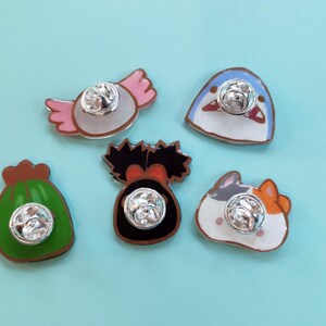 May include: Five enamel pins with different designs. The pins are on a light blue background. The designs include a green bag, a pink and white candy, a blue fish, a black and white creature, and a white cat with orange and black markings.