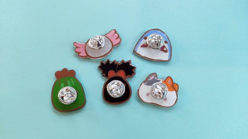 May include: Five enamel pins with different designs. The pins are on a light blue background. The designs include a green bag, a pink and white candy, a blue fish, a black and white creature, and a white cat with orange and black markings.