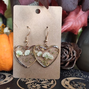 May include: A pair of gold heart-shaped earrings with white pressed flowers and gold flakes inside the resin. The earrings are on a brown card.