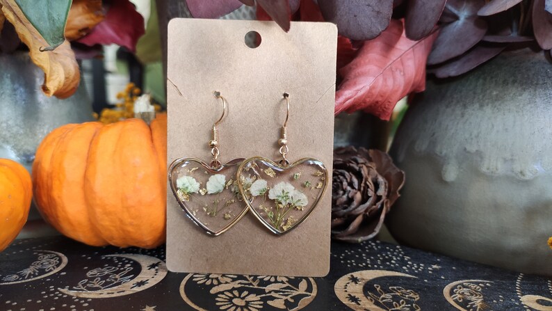 May include: A pair of gold heart-shaped earrings with white pressed flowers and gold flakes inside the resin. The earrings are on a brown card.