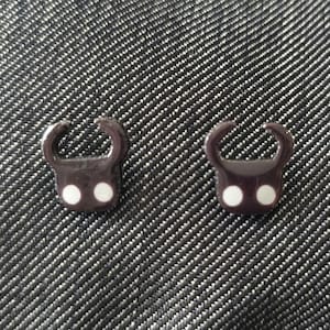 May include: Two dark brown, stylized charm pendants with a rounded top and two white dots. The pendants are placed on a textured, dark gray surface. The charms have a unique, abstract design.