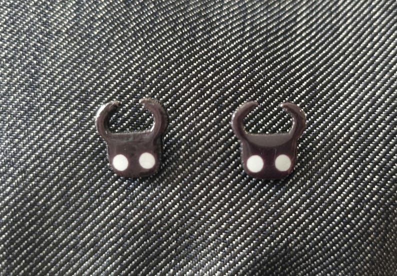 May include: Two dark brown, stylized charm pendants with a rounded top and two white dots. The pendants are placed on a textured, dark gray surface. The charms have a unique, abstract design.