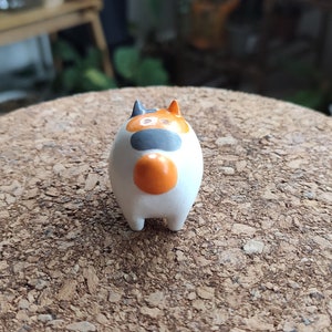 K&ouml;nnte beinhalten: Eine kleine, wei&szlig;e Keramikfigur eines Katers mit orangefarbenen, schwarzen und wei&szlig;en Markierungen. Die Katze sitzt auf einer Korkoberfl&auml;che.