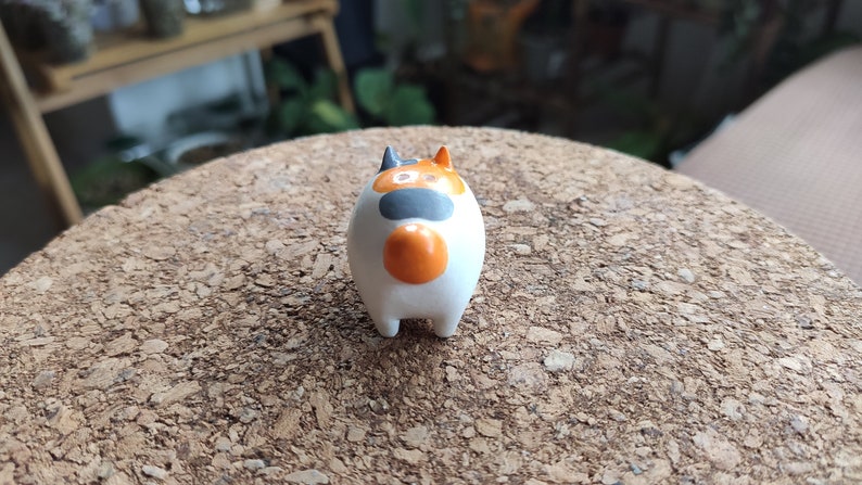 K&ouml;nnte beinhalten: Eine kleine, wei&szlig;e Keramikfigur eines Katers mit orangefarbenen, schwarzen und wei&szlig;en Markierungen. Die Katze sitzt auf einer Korkoberfl&auml;che.