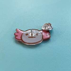 May include: A pink and white axolotl pin with a brown outline. The pin has a silver back and a safety clasp.