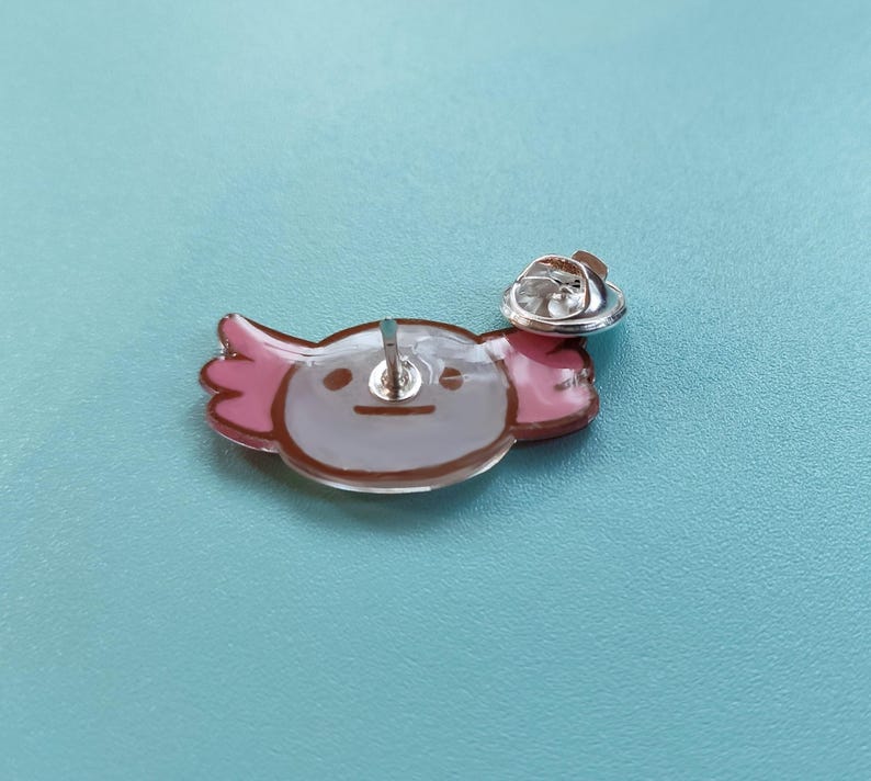 May include: A pink and white axolotl pin with a brown outline. The pin has a silver back and a safety clasp.