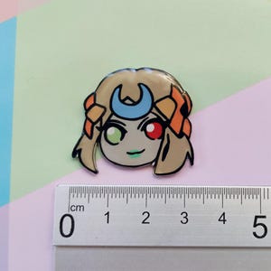 May include: A cartoon character with blonde hair, a crescent moon headband, and green and red eyes. The character is wearing a light orange and brown outfit. The character is approximately 2 cm tall.