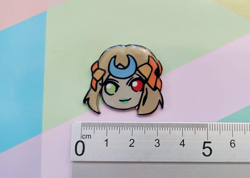 May include: A cartoon character with blonde hair, a crescent moon headband, and green and red eyes. The character is wearing a light orange and brown outfit. The character is approximately 2 cm tall.