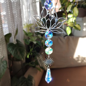 May include: A silver metal lotus flower suncatcher with iridescent blue and green crystal prisms hanging from it. The suncatcher is hanging from a chain and has a star-shaped crystal at the bottom.