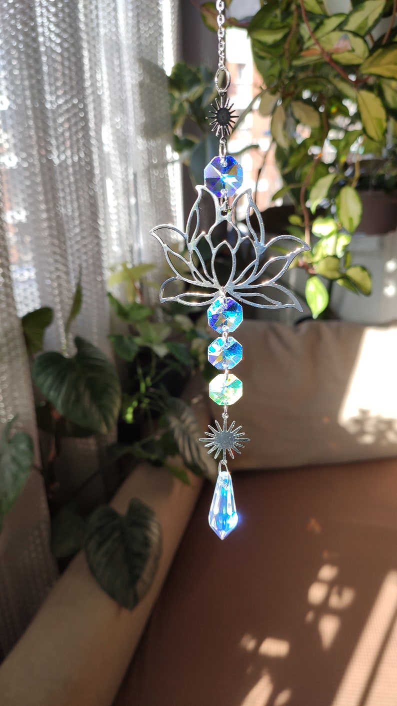 May include: A silver metal lotus flower suncatcher with iridescent blue and green crystal prisms hanging from it. The suncatcher is hanging from a chain and has a star-shaped crystal at the bottom.