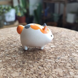 K&ouml;nnte beinhalten: Eine kleine, wei&szlig;e Keramikfigur eines Katers mit orangefarbenen und schwarzen Markierungen. Die Katze sitzt auf einer Korkoberfl&auml;che.