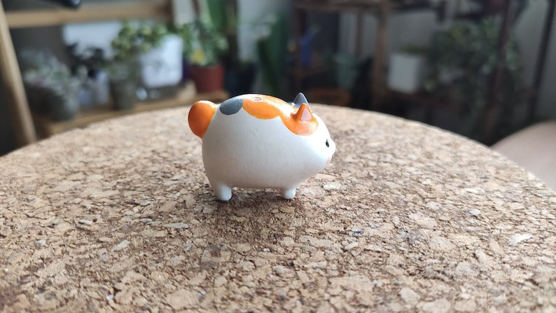 K&ouml;nnte beinhalten: Eine kleine, wei&szlig;e Keramikfigur eines Katers mit orangefarbenen und schwarzen Markierungen. Die Katze sitzt auf einer Korkoberfl&auml;che.