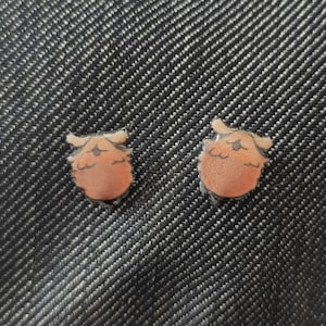 May include: Two small, orange and tan owl-shaped earrings. The earrings are set against a dark denim background. The owl design features a round body, small wings, and a detailed face.
