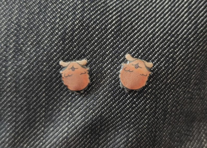 May include: Two small, orange and tan owl-shaped earrings. The earrings are set against a dark denim background. The owl design features a round body, small wings, and a detailed face.