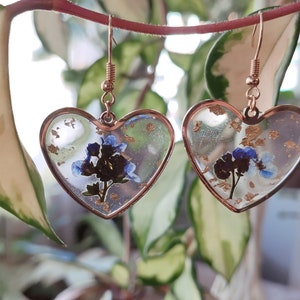 May include: A pair of heart-shaped earrings with a gold frame and blue flowers pressed inside. The earrings have a gold-colored hook.