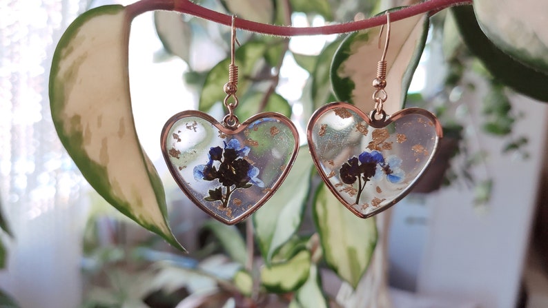 May include: A pair of heart-shaped earrings with a gold frame and blue flowers pressed inside. The earrings have a gold-colored hook.