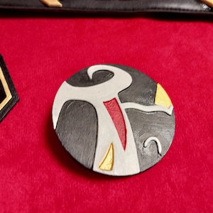 May include: A round, black, white, gold, and red leather patch with a stylized design. The design features a white figure with a red stripe and gold accents.
