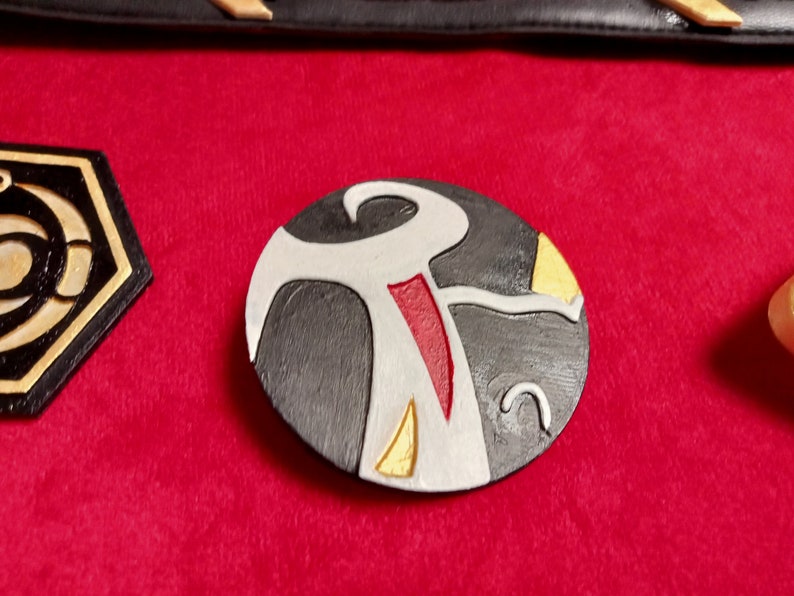 May include: A round, black, white, gold, and red leather patch with a stylized design. The design features a white figure with a red stripe and gold accents.