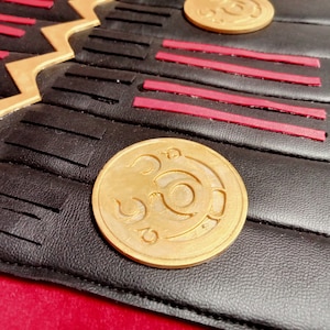 May include: Close-up of a black leather surface with a gold-colored circular design. The design features a stylized symbol with a circular shape and a curved line.