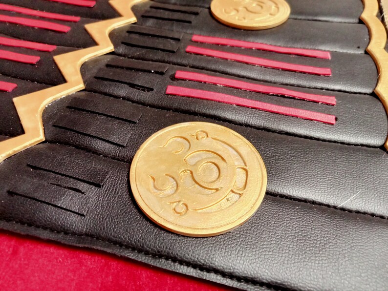 May include: Close-up of a black leather surface with a gold-colored circular design. The design features a stylized symbol with a circular shape and a curved line.
