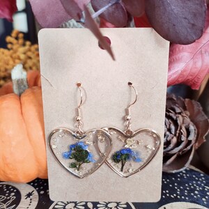 May include: A pair of gold heart-shaped earrings with blue and green pressed flowers and gold flakes inside the resin.