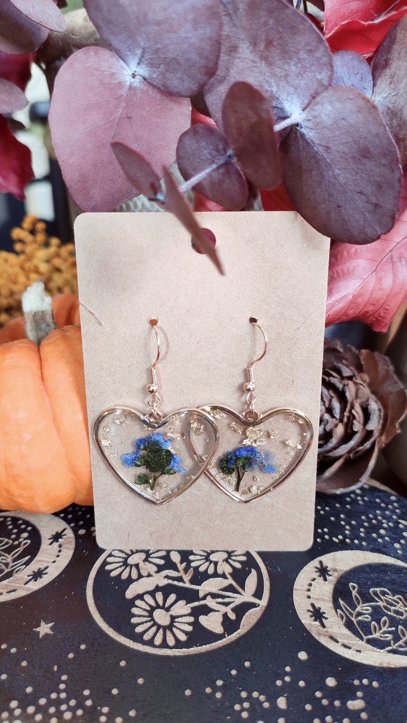 May include: A pair of gold heart-shaped earrings with blue and green pressed flowers and gold flakes inside the resin.