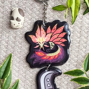 May include: A black keychain with a colorful phoenix design and a silver keyring. The keychain also features a second charm with a helmet design. A small white and gray cat figurine is also visible.