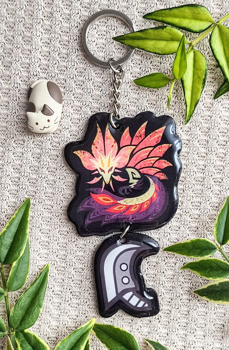 May include: A black keychain with a colorful phoenix design and a silver keyring. The keychain also features a second charm with a helmet design. A small white and gray cat figurine is also visible.