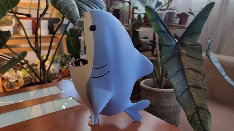 May include: A light blue shark figurine with a white belly and open mouth, showcasing sharp teeth and a red interior. The cartoonish shark has black eyes and is standing on a wooden surface. The background features lush green plants.