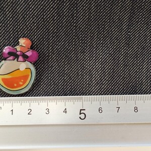May include: A small, round, clear glass bottle with a pink bow and orange liquid inside. The bottle is about 2 cm in diameter.