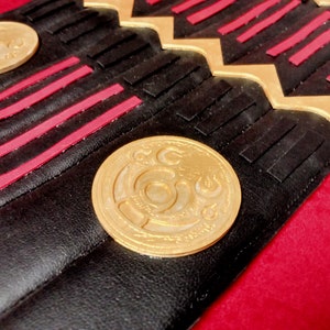 May include: Close-up of a black leather belt with gold and red accents. The belt has a repeating pattern of gold and red stripes and a gold coin with a decorative design.