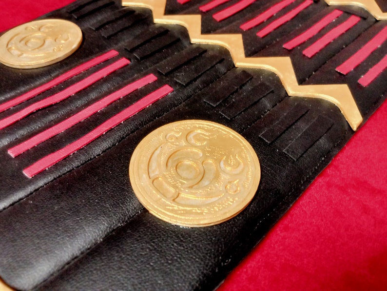 May include: Close-up of a black leather belt with gold and red accents. The belt has a repeating pattern of gold and red stripes and a gold coin with a decorative design.