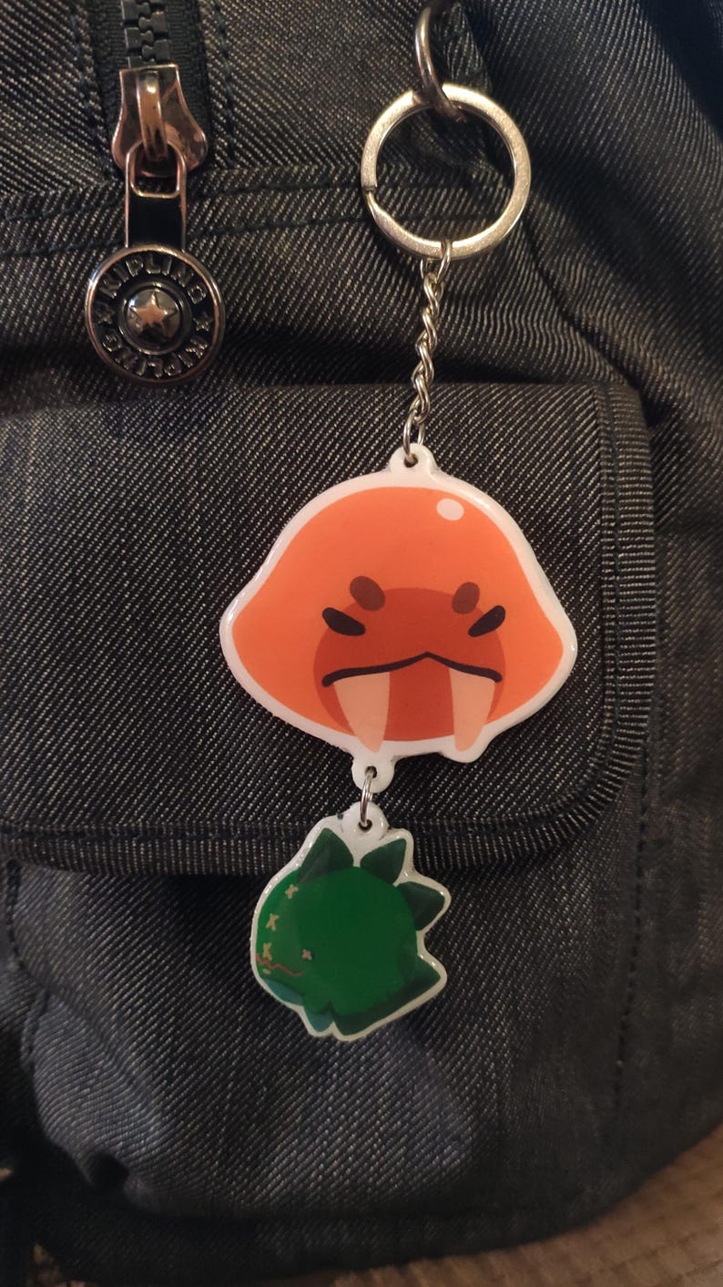 Puede incluir: Un llavero plateado con un personaje de dibujos animados rojo y naranja con dientes grandes y un personaje de dibujos animados verde con una estrella blanca en la cabeza.