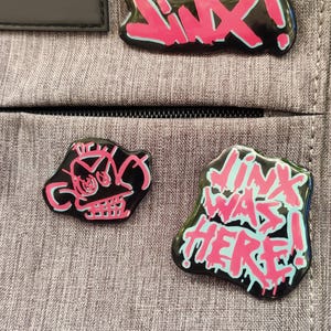 May include: Three black enamel pins with pink and blue accents. The pins feature a skull, the word "JINX!" and the phrase "LINK WAS HERE!"
