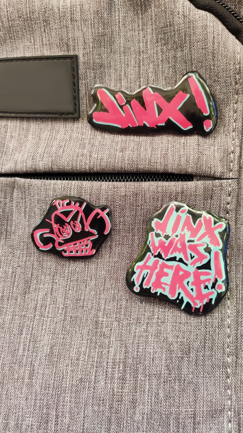 May include: Three black enamel pins with pink and blue accents. The pins feature a skull, the word "JINX!" and the phrase "LINK WAS HERE!"