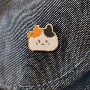 May include: A white enamel pin with a cartoon cat design. The cat has orange and black fur and a white face with black eyes and a pink nose.