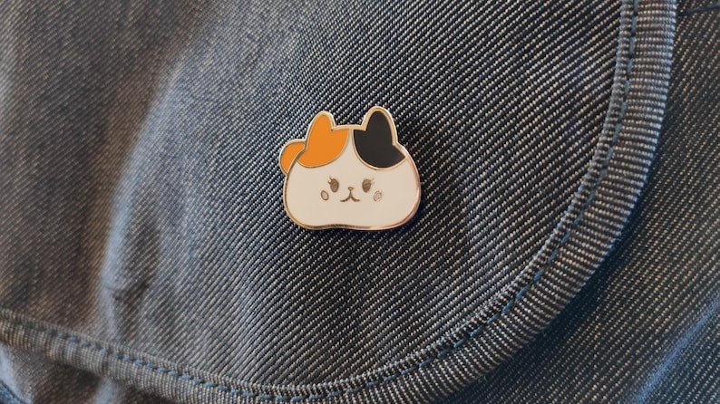 May include: A white enamel pin with a cartoon cat design. The cat has orange and black fur and a white face with black eyes and a pink nose.
