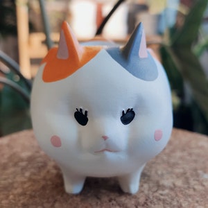 Puede incluir: Un macetero de cer&aacute;mica blanca con forma de gato con detalles en naranja, gris y rosa. El gato tiene ojos negros y una nariz rosa.