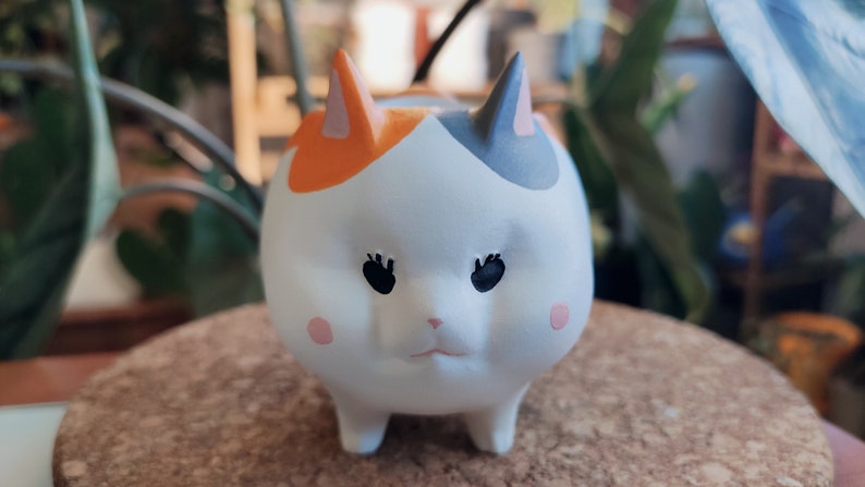 Puede incluir: Un macetero de cer&aacute;mica blanca con forma de gato con detalles en naranja, gris y rosa. El gato tiene ojos negros y una nariz rosa.
