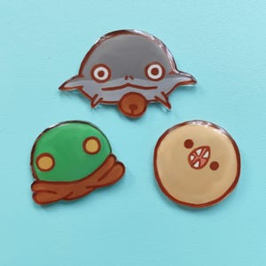 May include: Three cartoon character pins. A gray alien-like creature with large eyes, a green creature with yellow eyes, and a tan creature with a red mouth.