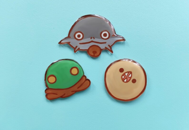 May include: Three cartoon character pins. A gray alien-like creature with large eyes, a green creature with yellow eyes, and a tan creature with a red mouth.