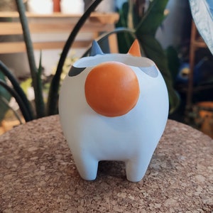 Puede incluir: Maceta de cer&aacute;mica blanca con forma de gato con detalles en negro y naranja. La maceta tiene una nariz redonda naranja y cuatro patas cortas.