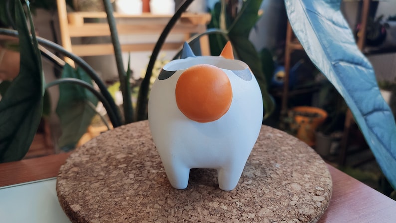 Puede incluir: Maceta de cer&aacute;mica blanca con forma de gato con detalles en negro y naranja. La maceta tiene una nariz redonda naranja y cuatro patas cortas.