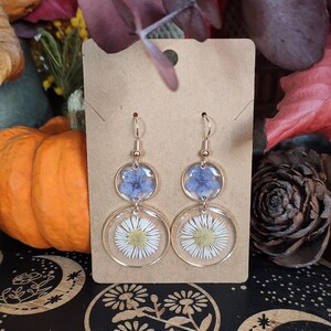 May include: Gold-coloured drop earrings with clear resin circles. Each earring has a blue flower in the top circle and a white daisy in the bottom. The earrings are displayed on a brown card, with a pumpkin and autumn leaves in the background.