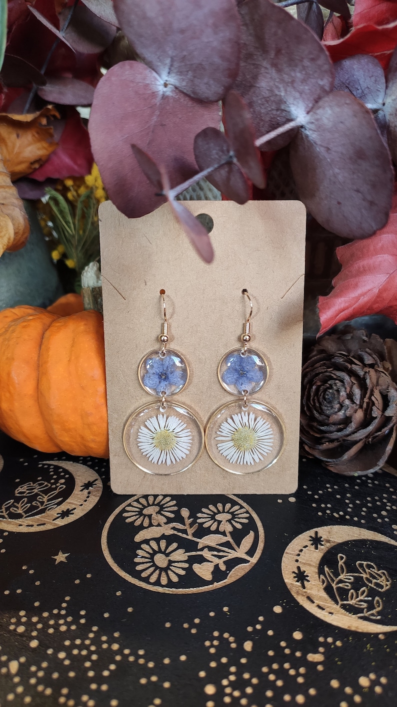May include: Gold-coloured drop earrings with clear resin circles. Each earring has a blue flower in the top circle and a white daisy in the bottom. The earrings are displayed on a brown card, with a pumpkin and autumn leaves in the background.