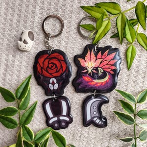 May include: Keychains with unique designs. One keychain features a red rose, another a stylized bird with pink and orange feathers. Two additional keychains have abstract shapes. A small cat figurine is also present.