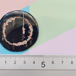 May include: A black and white button with a red and white design. The button is 5 cm in diameter.