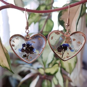 May include: A pair of gold heart-shaped earrings with blue and white pressed flowers and gold flakes inside the resin.